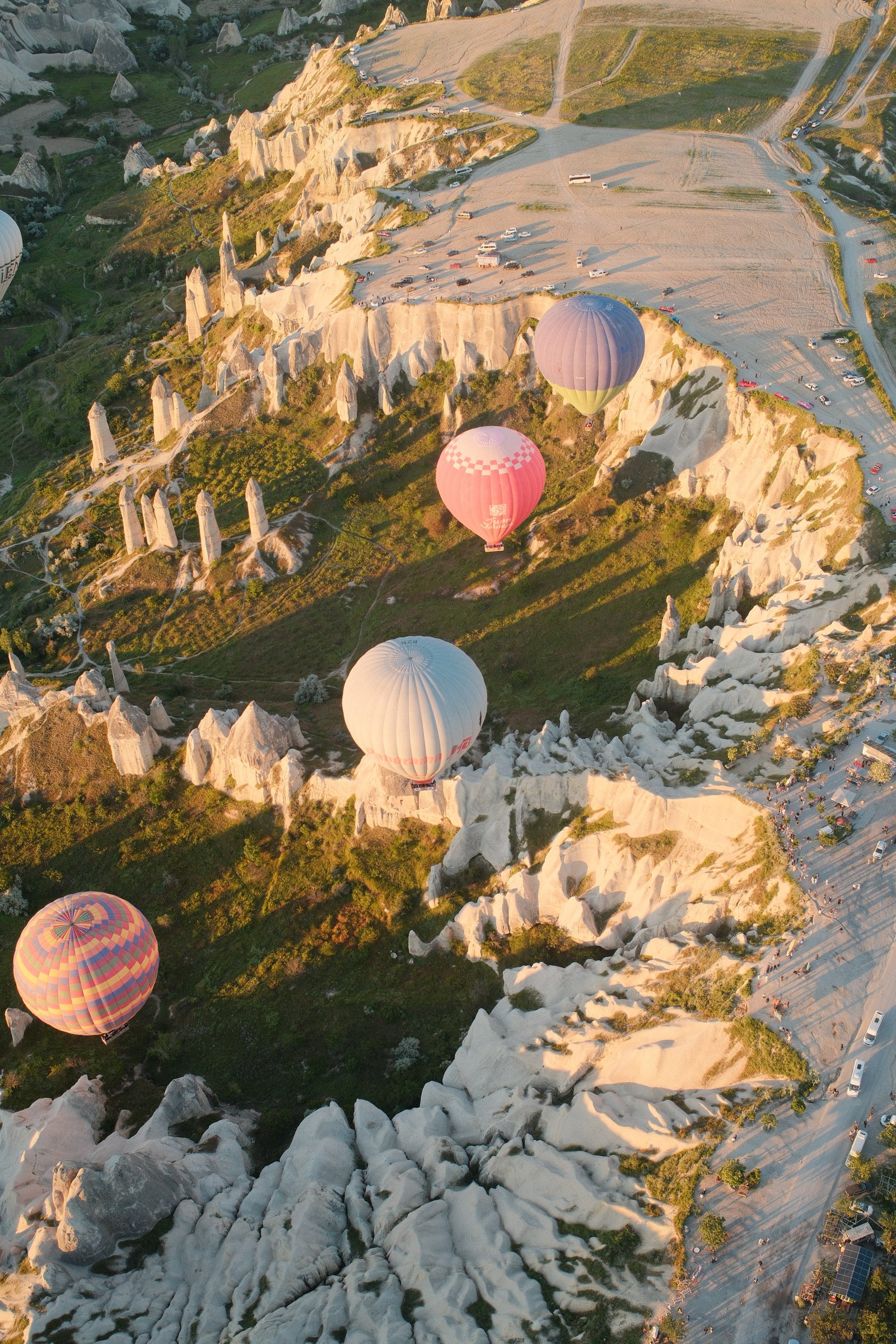 Several balloons descending into the valley among the fairy chimneys, cars and onlookers gathered below.