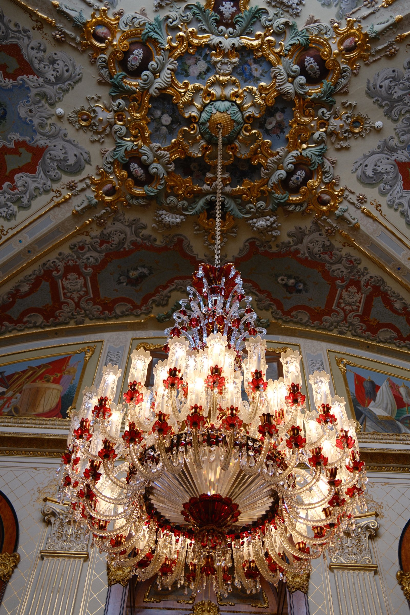 A large red and crystal chandelier hanging from a painted, gilded ceiling.