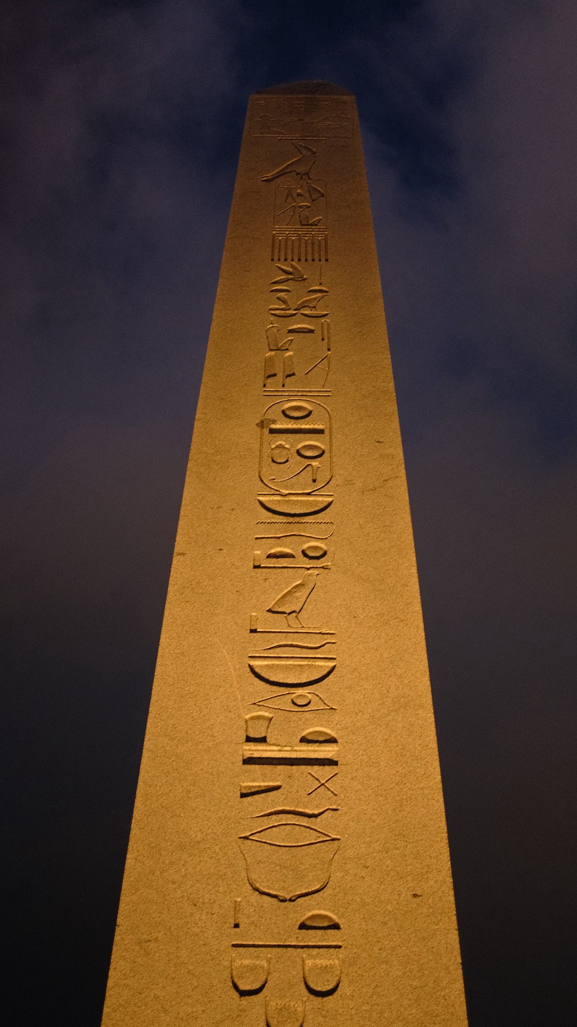 An ancient stone obelisk covered in hieroglyphic carvings, floodlit against the night sky.