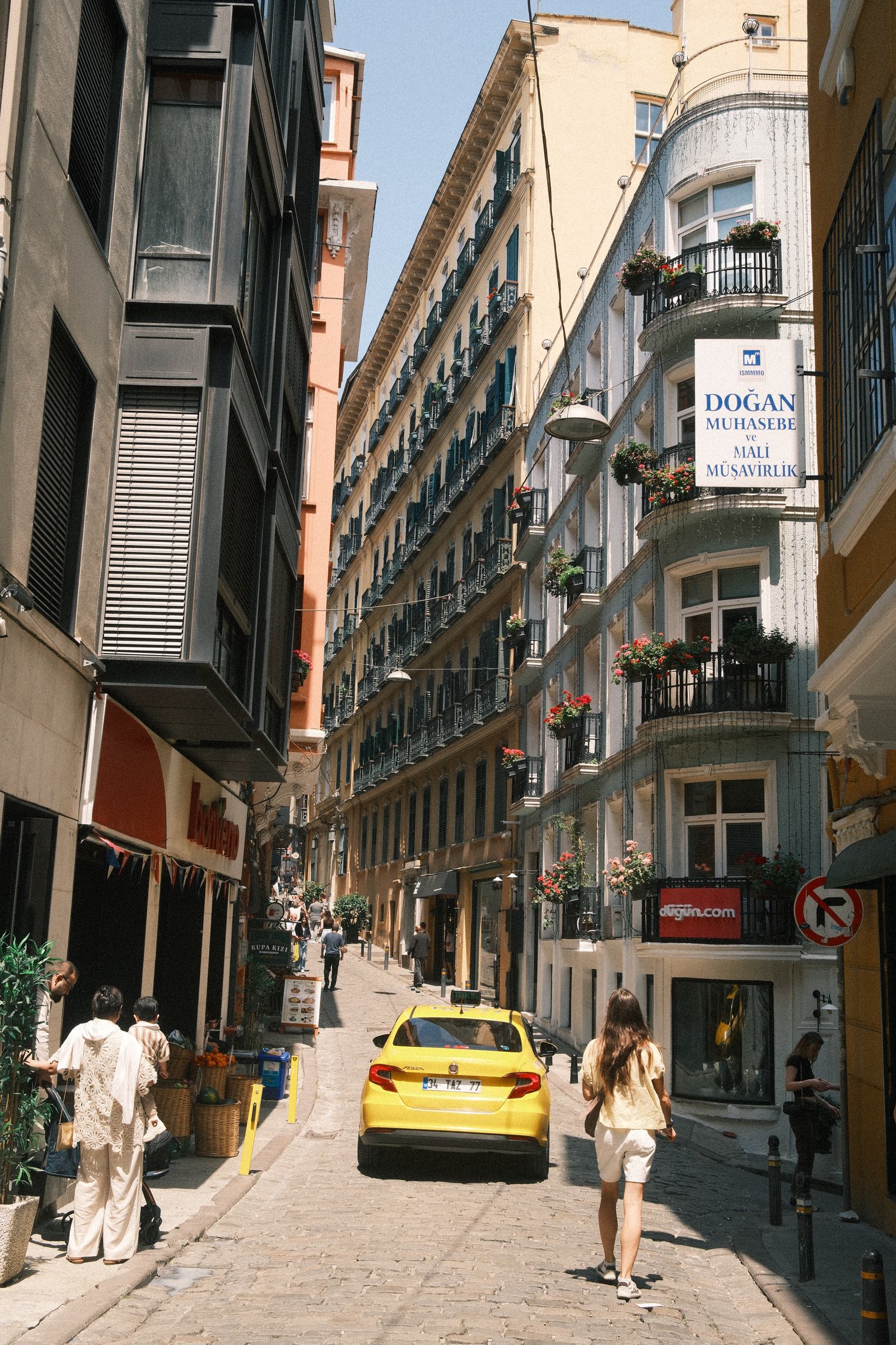 A narrow street lined with balconied apartment buildings, a yellow taxi and pedestrians passing through.