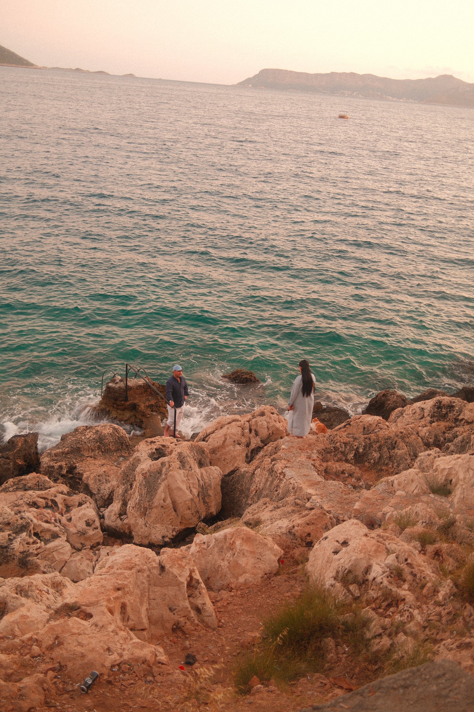 Two people and a dog on the rocky Kas coastline, looking out at the sea at dusk.