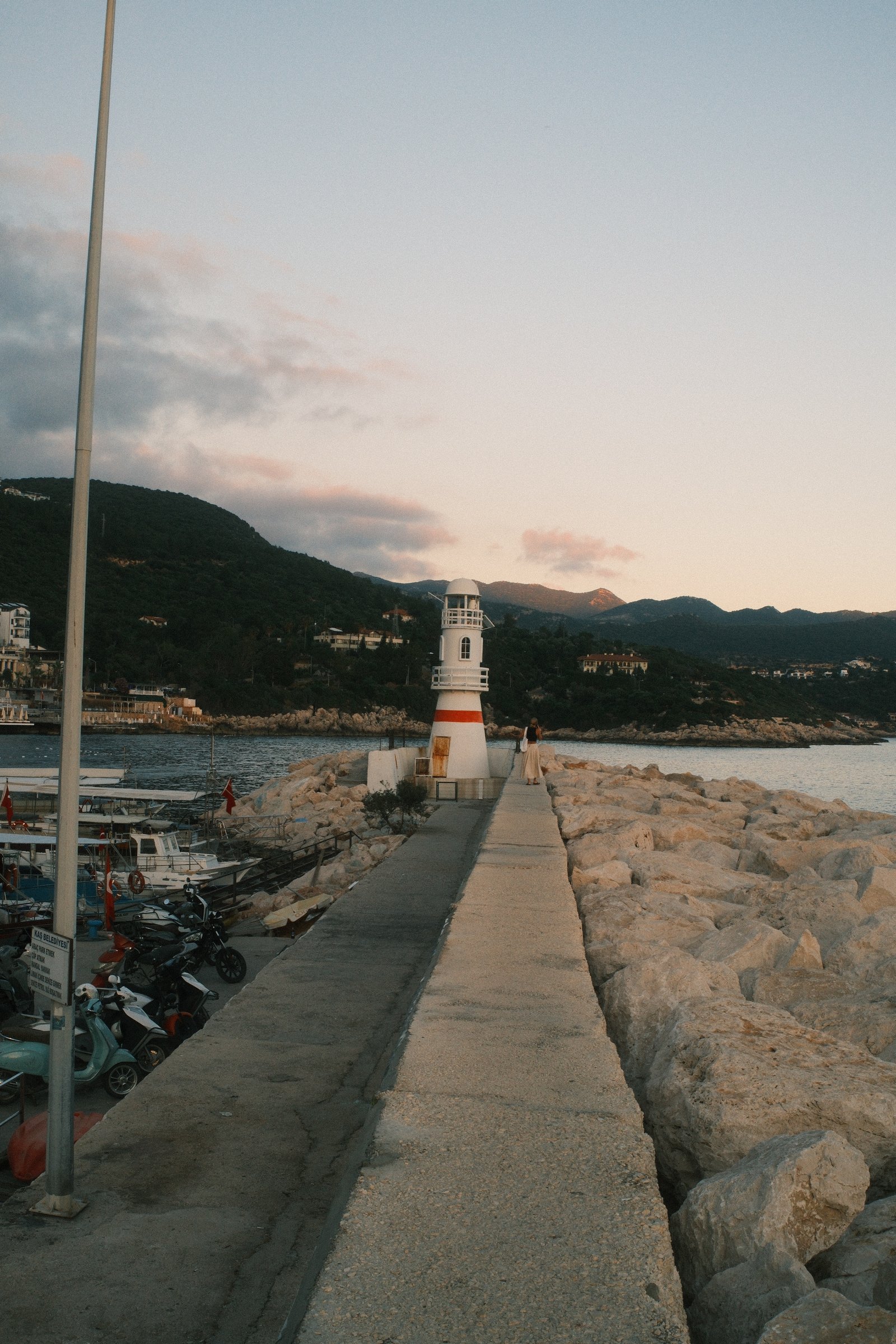 The same lighthouse from the pier, a woman walking toward it as the sky turns pink.
