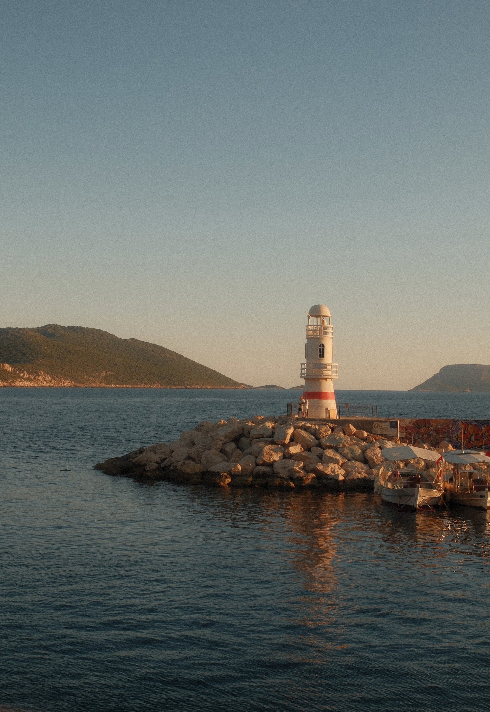 A red and white lighthouse at the end of a rocky breakwater, boats moored beside it at sunset.