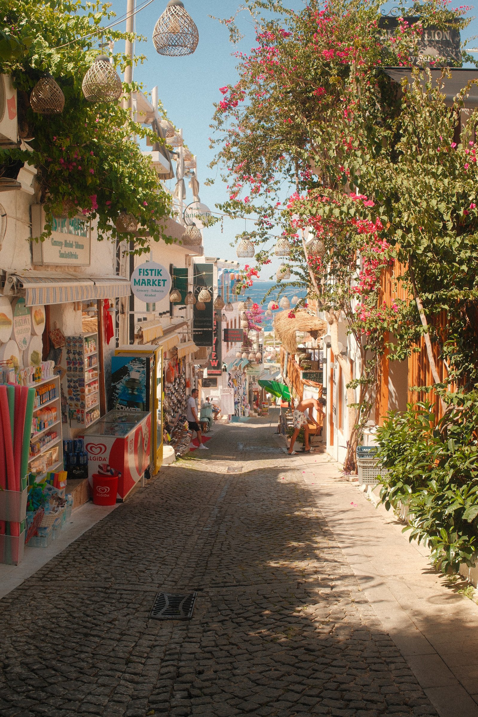 A cobblestone lane through Kas lined with shops, hanging lamps, and bougainvillea, the sea visible ahead.