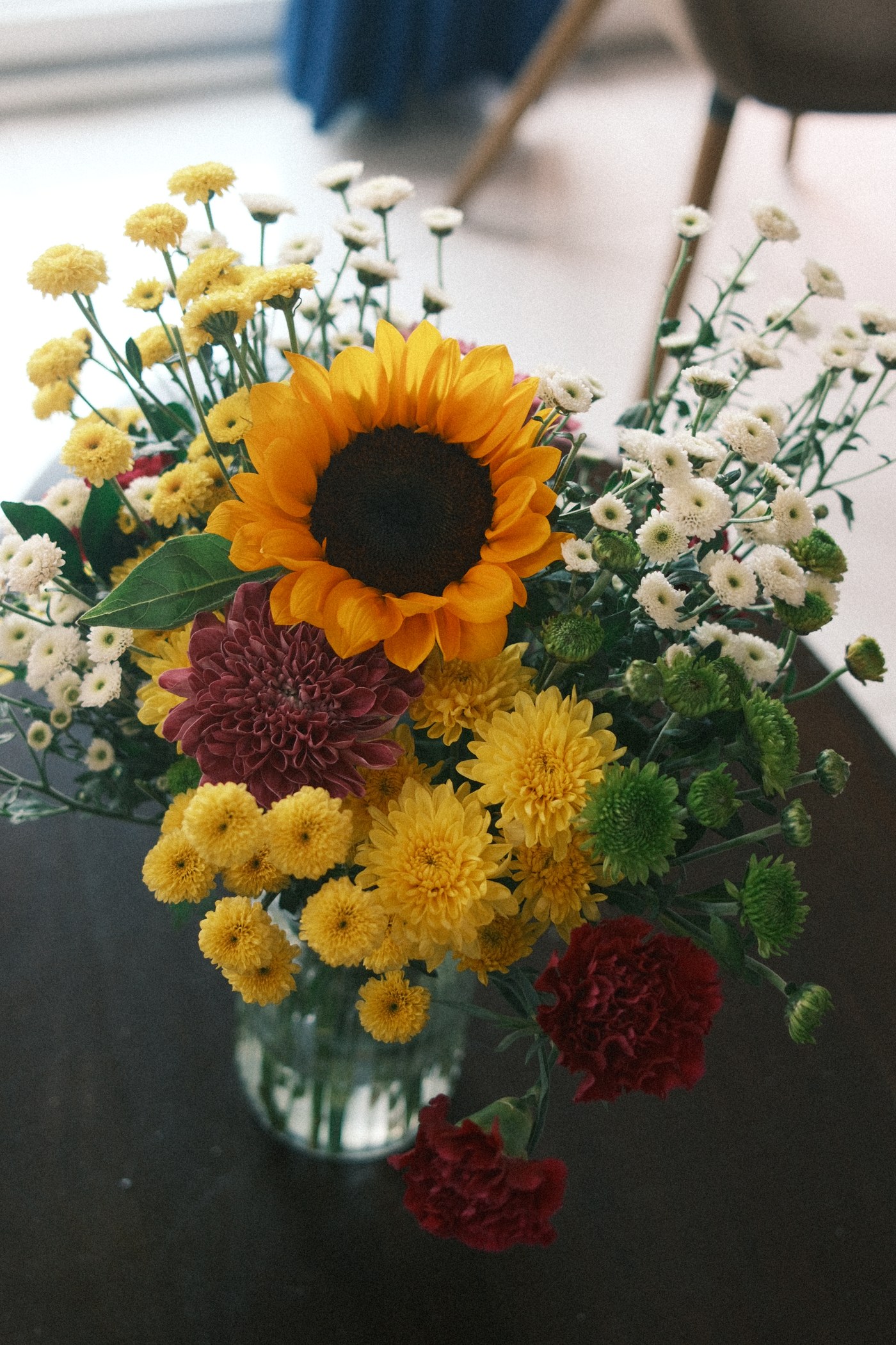 A bouquet of sunflowers, chrysanthemums, and carnations in a glass vase, shot from above against a dark background.